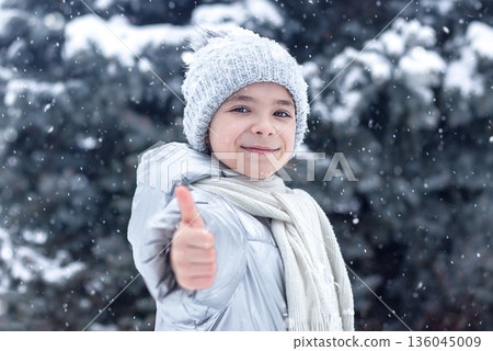 Smiling girl in winter gear gives thumbs up as snow falls in a snowy forest Smiling girl in winter gear gives thumbs up as snow falls in a snowy forest 136045009