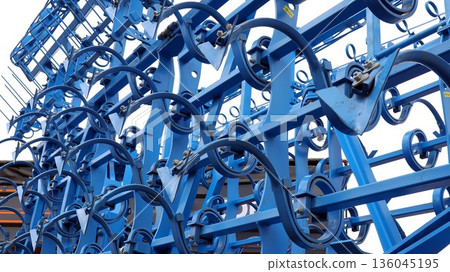 A modern blue metal agricultural implement is positioned in a farm area. This tool is designed for soil preparation, likely serving as a spring tine harrow or cultivator. A modern blue metal agricultural implement is positioned in a farm area. This tool is designed for soil preparation, likely serving as a spring tine harrow or cultivator. 136045195