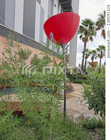 A large red flower-shaped lantern is placed near a shopping center among green plants. It adds color to the area and attracts attention from visitors passing by. 136045328