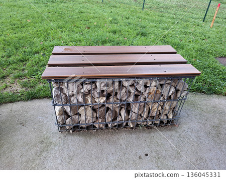 A park bench is placed in a parking lot alongside a busy highway. It is made of metal mesh and filled with stones for support and design. People can sit and take a break here. A park bench is placed in a parking lot alongside a busy highway. It is made of metal mesh and filled with stones for support and design. People can sit and take a break here. 136045331