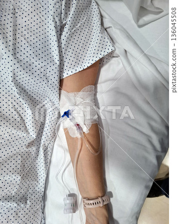 A close up of a patient's arm with an intravenous drip. The arm is covered in a bandage, and medical equipment is attached. This happens in a hospital setting. 136045508