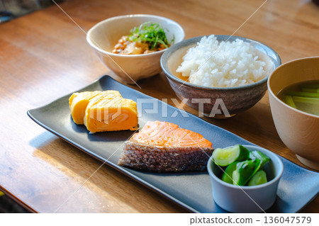 一份日式早餐套餐（烤鮭魚、玉子燒、味噌湯、白飯和一份小菜）擺放在沐浴著晨光的木桌上。 136047579