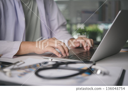Male physician using computer for online patient consultation, managing digital health records, medical diagnosis on desk with stethoscope, representing telehealth and modern healthcare services. Male physician using computer for online patient consultation, managing digital health records, medical diagnosis on desk with stethoscope, representing telehealth and modern healthcare services. 136048754
