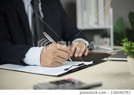 Businessman in suit signing contract document at office desk. Closeup hand holding pen for business agreement, partnership deal, legal commitment, finance investment, or insurance policy approval. Businessman in suit signing contract document at office desk. Closeup hand holding pen for business agreement, partnership deal, legal commitment, finance investment, or insurance policy approval. 136048761