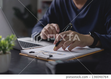 Businessman analyzing financial data report, hand with pen pointing at chart. Man working with laptop office desk, reviewing business performance, investment growth, and market strategy for success. Businessman analyzing financial data report, hand with pen pointing at chart. Man working with laptop office desk, reviewing business performance, investment growth, and market strategy for success. 136048764