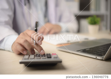 Doctor calculates medical billing at office desk with calculator laptop paperwork for insurance claim cost control finance management healthcare administration concept 136048778