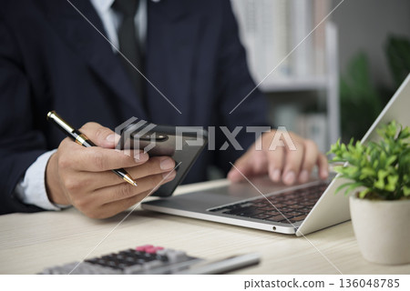 Manager checks phone while typing on laptop in office for finance report email communication mobile banking productivity technology workflow remote work concept modern business. 136048785