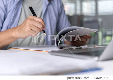 Office worker reviews document, paperwork with pen, writing notes beside laptop on desk to analyze report and organize tasks. close-up hands convey accuracy, focus, planning, and productivity concept. 136049040
