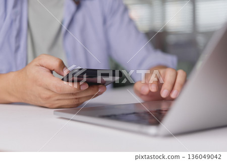 Freelancer uses smartphone, phone, and mobile while typing on laptop computer in home office to work online and enhance communication; detailed hands illustrate technology and productivity. 136049042