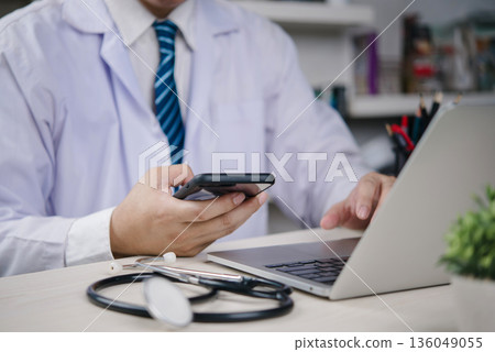 Healthcare medical Doctor with stethoscope works on laptop while checking smartphone to manage EHR records, telehealth messages, and patient data securely. concept connected care and compliance. 136049055