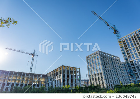Workers and cranes are busy at a construction site, bringing a new building to life under the bright blue sky and warm sunshine 136049742