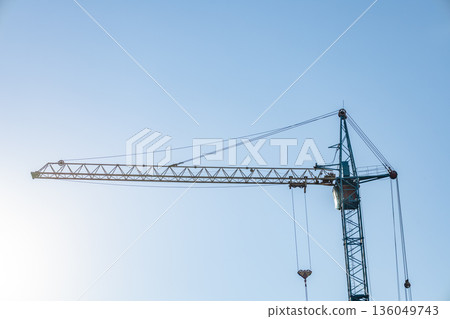 A tall construction crane stands against a clear blue sky, ready to lift materials at a lively construction site under bright sunlight A tall construction crane stands against a clear blue sky, ready to lift materials at a lively construction site under bright sunlight 136049743
