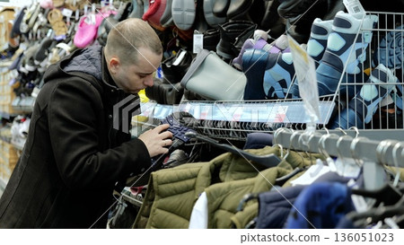 Male customer carefully examining trekking shoe sole, inspecting quality and materials while browsing sporting goods store footwear section 136051023