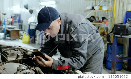 Auto mechanic using a smartphone app to consult repair instructions while working on a car engine inside a professional workshop, troubleshooting and fixing mechanical issues Auto mechanic using a smartphone app to consult repair instructions while working on a car engine inside a professional workshop, troubleshooting and fixing mechanical issues 136051032