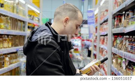 Focused shopper examining olive oil bottle label, selecting quality grocery item for household needs during supermarket trip Focused shopper examining olive oil bottle label, selecting quality grocery item for household needs during supermarket trip 136051037