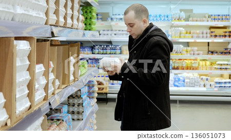 Male shopper checking egg carton packaging, scanning nutrition details in refrigerated grocery section, comparing quality for nutritious dietary selection 136051073