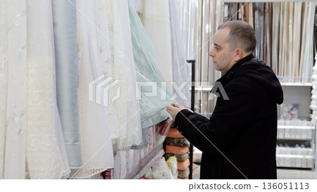 Customer carefully examining curtain samples hanging on display rack in home improvement store, selecting window treatment for interior design project 136051113