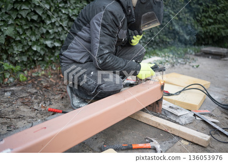 Construction worker in protective mask and gloves welding a steel beam outdoors, bright sparks and smoke. Metalwork, fabrication and repair concept. Construction worker in protective mask and gloves welding a steel beam outdoors, bright sparks and smoke. Metalwork, fabrication and repair concept. 136053976