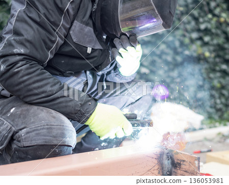 Construction worker in protective mask and gloves welding a steel beam outdoors, bright sparks and smoke. Metalwork, fabrication and repair concept. Construction worker in protective mask and gloves welding a steel beam outdoors, bright sparks and smoke. Metalwork, fabrication and repair concept. 136053981