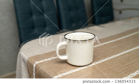 White enamel mug on patterned tablecloth in cozy kitchen setting White enamel mug on patterned tablecloth in cozy kitchen setting 136054315