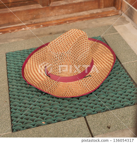 Woven straw hat with red trim on green doormat near wooden door Woven straw hat with red trim on green doormat near wooden door 136054317