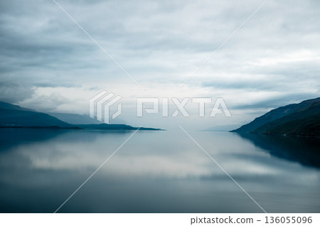 Calm fjord lake with misty mountains and dramatic cloudy sky in Norway Calm fjord lake with misty mountains and dramatic cloudy sky in Norway 136055096