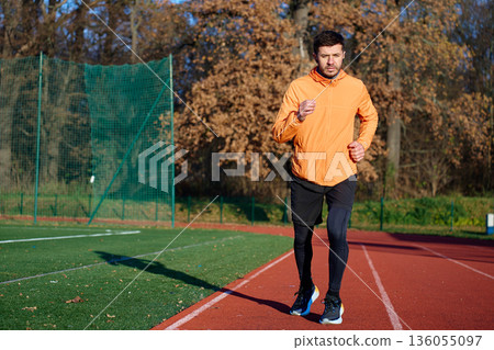 Man in orange sports jacket jogging on outdoor running track at stadium Man in orange sports jacket jogging on outdoor running track at stadium 136055097