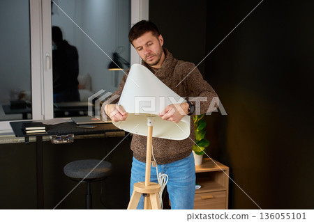 Man standing in home interior installing white lampshade on floor lamp during furniture assembly 136055101