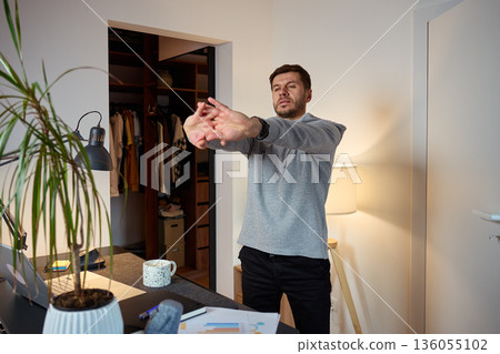 Man standing at home office desk stretching arms forward during work break Man standing at home office desk stretching arms forward during work break 136055102