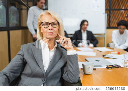 Portrait of female team leader businesswoman in office, colleagues meeting in background. Smiling confident business leader woman looking at camera sitting in office. Boss leader professional worker 136055288