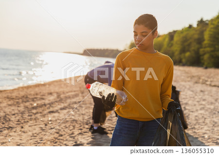 Earth day. Volunteers activists team collects garbage cleaning of beach coastal zone. Woman puts plastic bottle trash in garbage bag on ocean shore. Environmental conservation coastal zone cleaning Earth day. Volunteers activists team collects garbage cleaning of beach coastal zone. Woman puts plastic bottle trash in garbage bag on ocean shore. Environmental conservation coastal zone cleaning 136055310