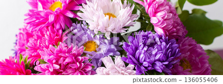 A group of flowers in shades of pink, purple, and white are gathered in a vase. The vase rests on a table. The setting is bright and shows the flowers clearly, banner 136055406