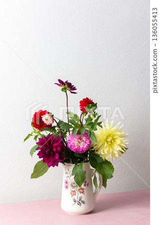 A vase holds a variety of fresh flowers with different colors. The flowers are placed on a pink table. Behind the vase is a plain wall in a bright room A vase holds a variety of fresh flowers with different colors. The flowers are placed on a pink table. Behind the vase is a plain wall in a bright room 136055413
