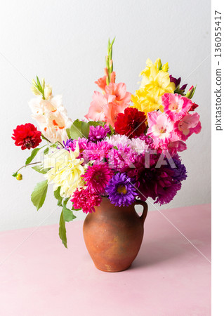 A clay vase holds a mix of flowers including glads and daisies. The vase is placed on a pink table. The room has bright light coming from outside A clay vase holds a mix of flowers including glads and daisies. The vase is placed on a pink table. The room has bright light coming from outside 136055417