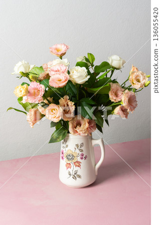 A bouquet of fresh flowers is arranged in a white pitcher on a pink surface. The flowers include pink and white blooms along with green leaves, seen in good light A bouquet of fresh flowers is arranged in a white pitcher on a pink surface. The flowers include pink and white blooms along with green leaves, seen in good light 136055420