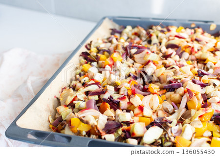 Fresh vegetables are chopped and placed on a baking tray ready for cooking in the oven. Colorful ingredients are showcased, and the setting is a welcoming kitchen 136055430