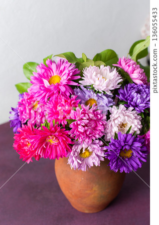 A clay pot holds a mix of colorful flowers arranged neatly on a pink table. The setting is bright with a plain background, showing natural beauty and simplicity A clay pot holds a mix of colorful flowers arranged neatly on a pink table. The setting is bright with a plain background, showing natural beauty and simplicity 136055433