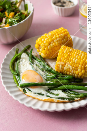 A healthy breakfast is served on a pink table. There are plates with asparagus, a fried egg, corn, and a fresh salad. Grapes and sunflower seeds are on the side A healthy breakfast is served on a pink table. There are plates with asparagus, a fried egg, corn, and a fresh salad. Grapes and sunflower seeds are on the side 136055434