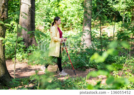 Middle-aged woman doing Nordic walking with special poles on a forest trail, promoting health, fitness, and connection with nature 136055452