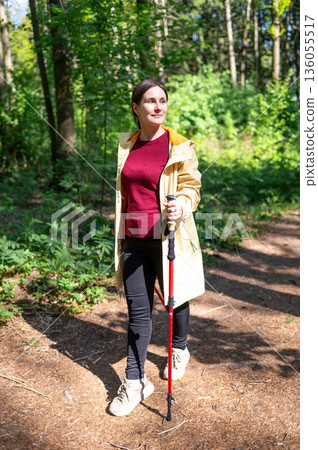 Young woman walking through a sunny forest, practicing Nordic walking with pole 136055517