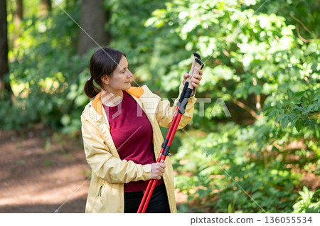 Woman on a forest walk examining her Nordic walking poles, enjoying nature and preparing for an active outdoor exercise Woman on a forest walk examining her Nordic walking poles, enjoying nature and preparing for an active outdoor exercise 136055534