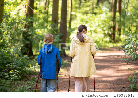 Rear view of mother and 11-year-old son hiking with Nordic trekking poles through a sunny forest trail 136055615
