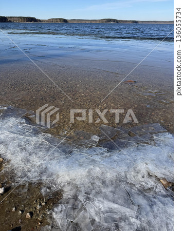 Cracked ice on a rocky shore of a thawed lake under a bright spring blue sky. A contrast of melting cold and warm seasonal renewal. Cracked ice on a rocky shore of a thawed lake under a bright spring blue sky. A contrast of melting cold and warm seasonal renewal. 136055734