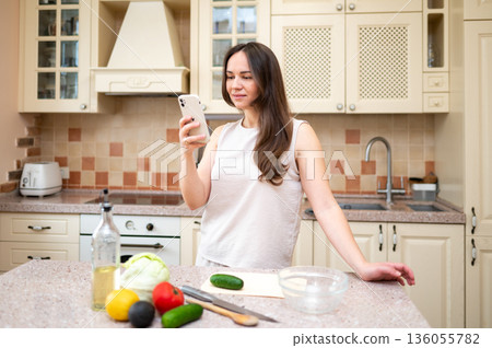 Smiling woman using smartphone while preparing fresh vegetable salad in cozy kitchen. Healthy lifestyle, home cooking and online recipe inspiration Smiling woman using smartphone while preparing fresh vegetable salad in cozy kitchen. Healthy lifestyle, home cooking and online recipe inspiration 136055782