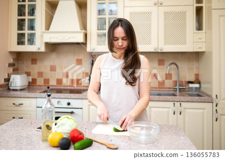 Smiling woman cutting cucumber for fresh vegetable salad in cozy kitchen. Healthy lifestyle, home cooking, diet and proper nutrition Smiling woman cutting cucumber for fresh vegetable salad in cozy kitchen. Healthy lifestyle, home cooking, diet and proper nutrition 136055783