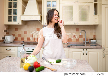 Young woman in bright kitchen eating cucumber and preparing salad. Fresh vegetables and ingredients on counter suggest healthy and balanced diet 136055785