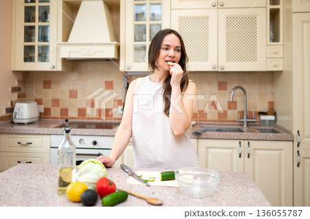 Young woman in bright kitchen eating cucumber and preparing salad. Fresh vegetables and ingredients on counter suggest healthy and balanced diet 136055787