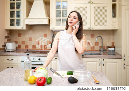 young woman in bright kitchen talking on phone and smiling, planning to cook salad. Fresh vegetables and board are on table 136055792
