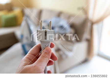 A plug with a power cord from a disconnected iron in a woman's hand against the background of an ironing board with an iron and a pile of crumpled laundry A plug with a power cord from a disconnected iron in a woman's hand against the background of an ironing board with an iron and a pile of crumpled laundry 136055802