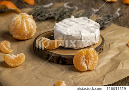 fresh white soft cheese with mold lies on a wooden board on crumpled yellow paper, soft round cheese and juicy sweet orange tangerines 136055859